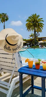Poolside lounge by palm trees with a sun hat reading “Hello sunshine” and colorful drinks on a blue table.
