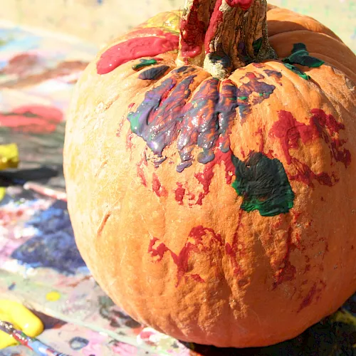 A painted orange pumpkin covered in colorful red, blue, green, and purple paint on a messy art-station table.