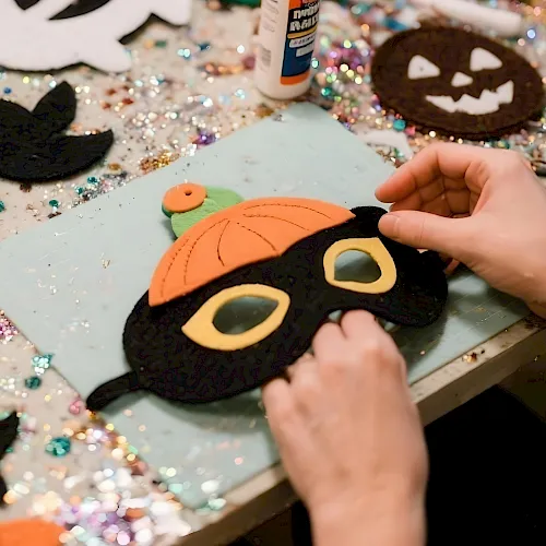 A person’s hands craft Halloween felt with a jack-o’-lantern face on a glittery table covered in decorations.